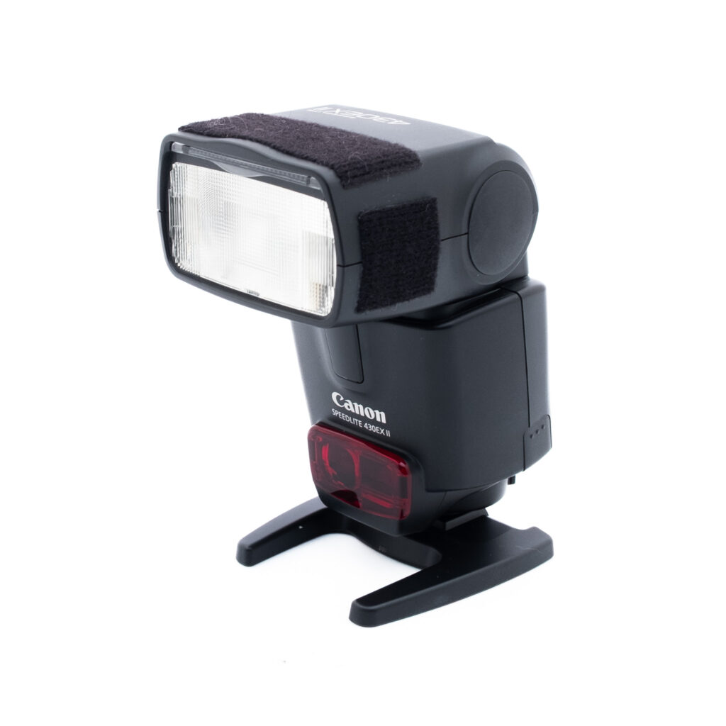 Canon 430EX II-2 Canon Speedlite 430EX II -kameran ulkoinen salama on kuvassa minijalustallaan valkoista taustaa vasten. Salamapäässä on diffuusiopaneeli ja tarranauhat päällä.