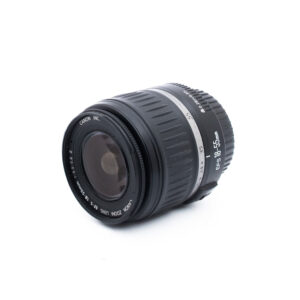 Musta Canon EF-S 18-55mm -kameran objektiivi on sijoitettu valkoiselle taustalle kulmaan, jossa näkyy sekä etulasi että sivumerkinnät.