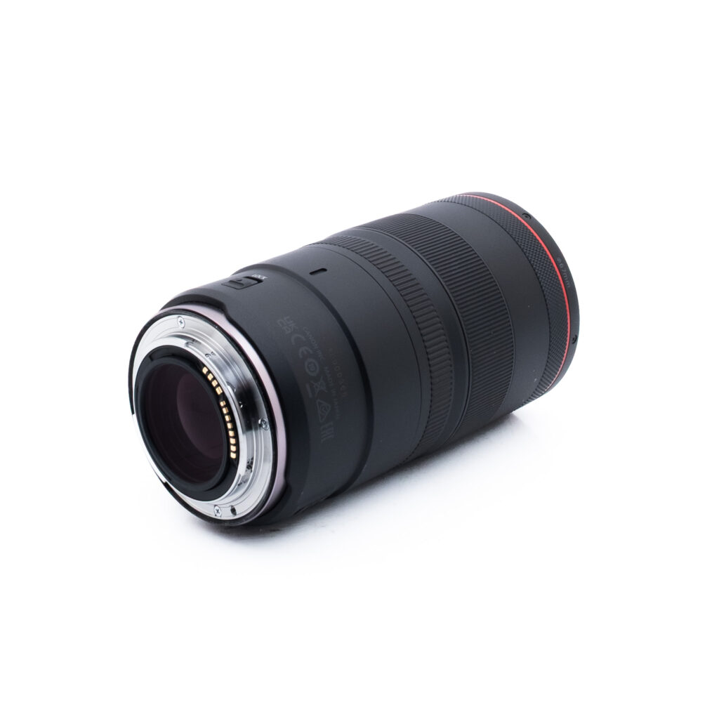 Canon RF 100mm f28 L Macro IS USM-1-2 Musta kameran objektiivi, jonka päässä on punainen rengas ja metalliset kiinnityskontaktit, valkoisella pohjalla.