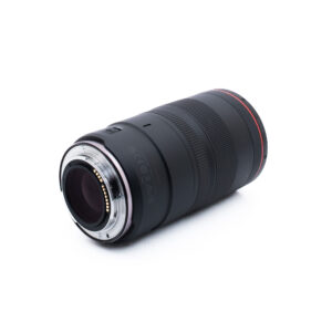 Canon RF 100mm f28 L Macro IS USM-1-2 Musta kameran objektiivi, jonka päässä on punainen rengas ja metalliset kiinnityskontaktit, valkoisella pohjalla.