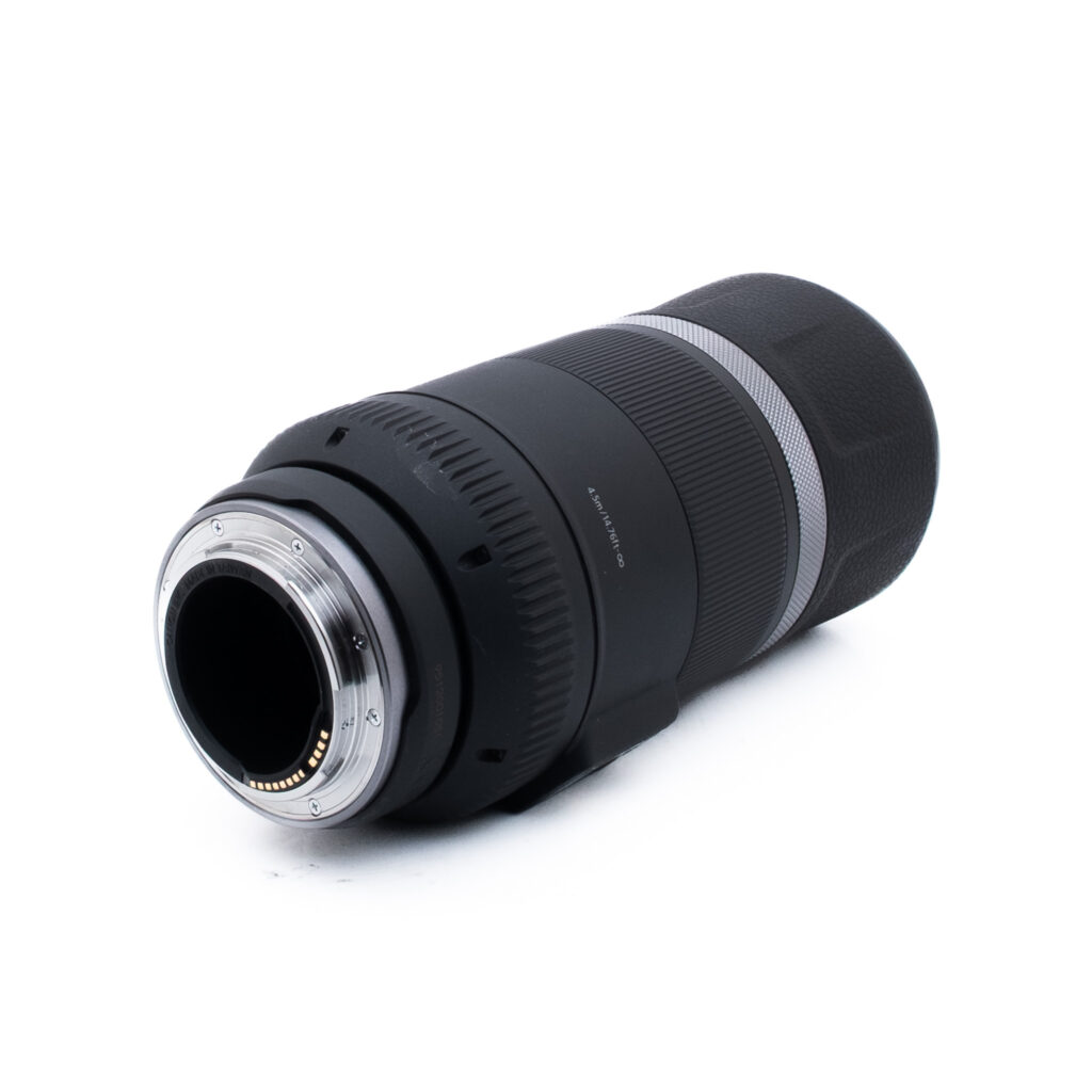 Canon RF 600mm-1-2 Musta kameran teleobjektiivi, jossa on hopeinen rengas pohjan lähellä, vinossa kuvattuna, jossa näkyy metallinen kiinnitys ja elektroniset koskettimet, valkoisella pohjalla.