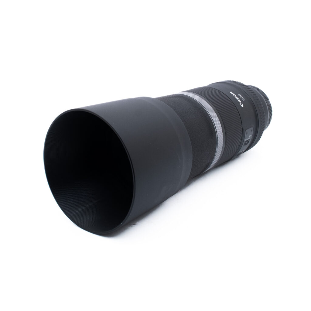 Canon RF 600mm-3-2 Musta Canon-telekameran objektiivi, jossa on suuri linssinsuojus ja joka on sijoitettu vinosti valkoiselle taustalle.