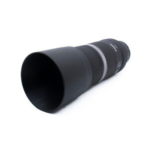 Canon RF 600mm-3-2 Musta Canon-telekameran objektiivi, jossa on suuri linssinsuojus ja joka on sijoitettu vinosti valkoiselle taustalle.