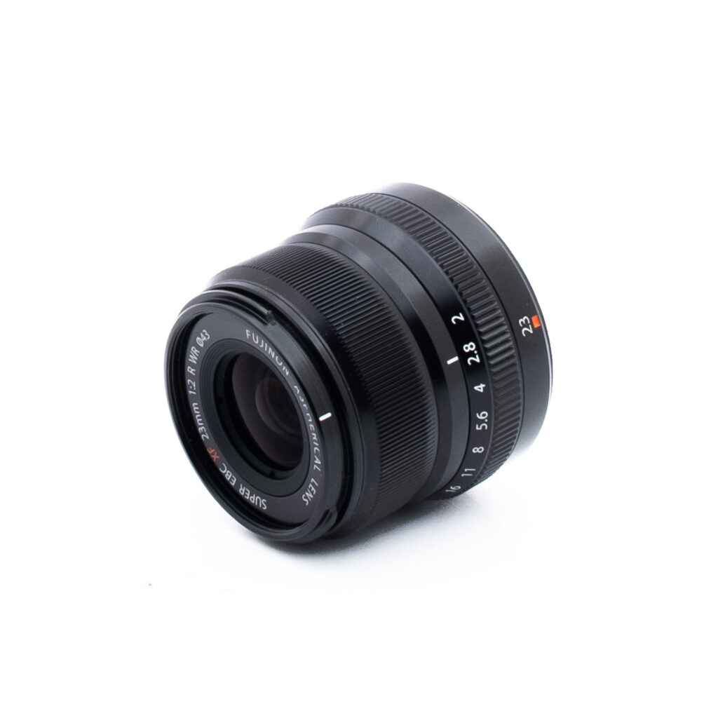 Fuji 23mm f2 R WR-2-2 Musta Fujinon-kameran objektiivi, jossa on valkoisia ja punaisia merkintöjä ja joka on sijoitettu vinoon valkoiselle taustalle. Aukko- ja tarkennusrenkaat sekä objektiivin tekniset tiedot ovat näkyvissä.