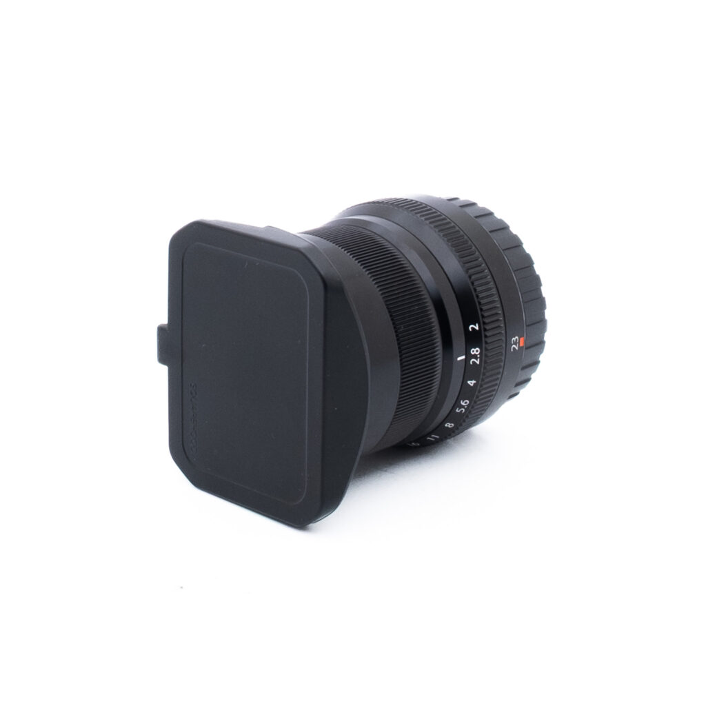 Fuji 23mm f2 R WR-3-2 Musta kameran objektiivi, johon on kiinnitetty neliönmuotoinen objektiivin suojus, valkoisella taustalla. Objektiivissa on harjattu tarkennusrengas ja aukon merkinnät sivulla.