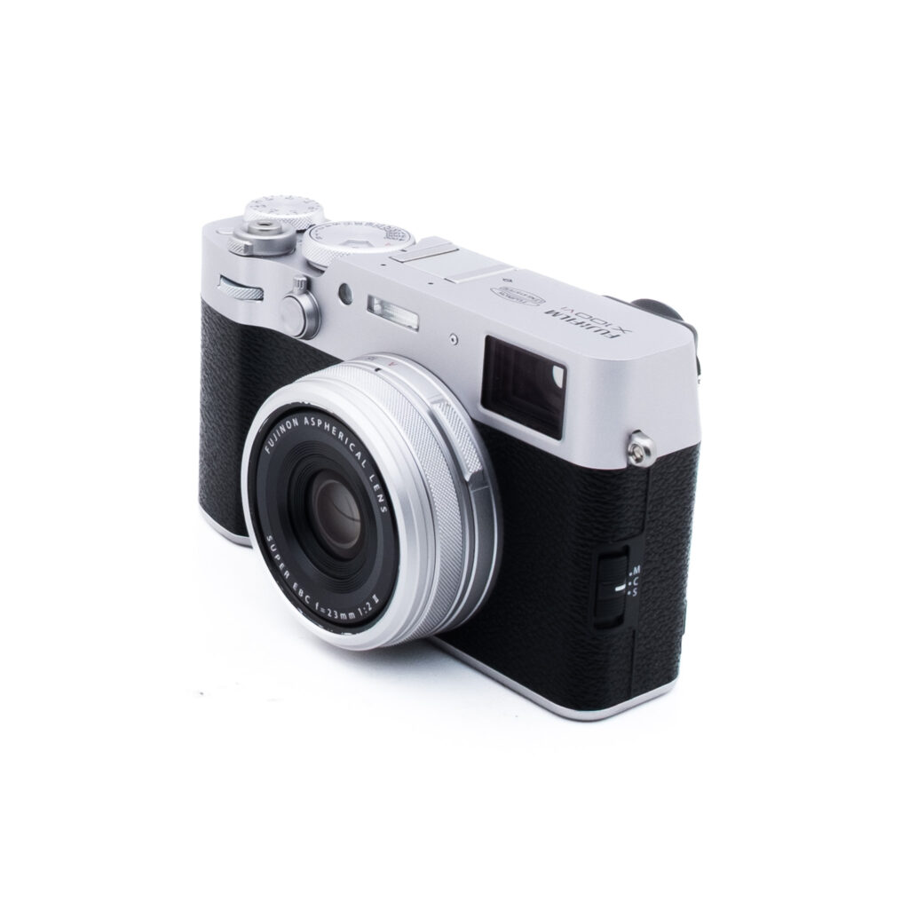 Fuji X100VI-4-3 Hopeanvärinen ja musta digitaalikamera, jossa on kiinteä linssi, on kallistettu hieman oikealle valkoisella taustalla. Kameran yläosassa on manuaaliset säätimet ja etupuolella on kuvioitu kahva.