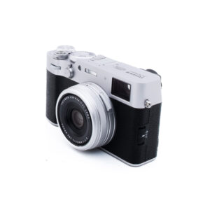 Fuji X100VI-4-3 Hopeanvärinen ja musta digitaalikamera, jossa on kiinteä linssi, on kallistettu hieman oikealle valkoisella taustalla. Kameran yläosassa on manuaaliset säätimet ja etupuolella on kuvioitu kahva.