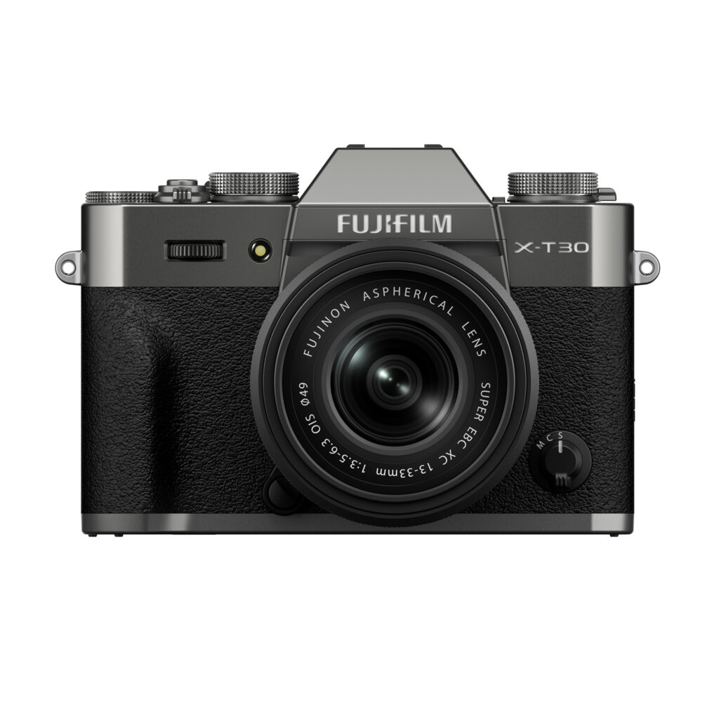 FujiX-T30IIIHarmaaLinssi1 Etukuva harmaasta Fujifilm X-T30 III -kamerasta, jossa on musta kuvioitu kahva ja kiinnitetty XC 13-33mm f/3.5-6.3 OIS -objektiivi, jossa on ylävalitsimet ja Fujifilmin tuotemerkki objektiivin yläpuolella.
