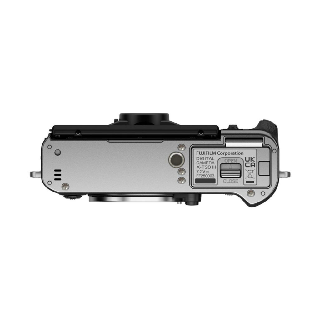 FujiX-T30IIIHopea5 Fujifilm X-T30 III Body Hopea -digitaalikameran hopeanvärinen ylhäältä alas -kuva, jossa näkyy akkulokero, tarrat ja osa objektiivista.