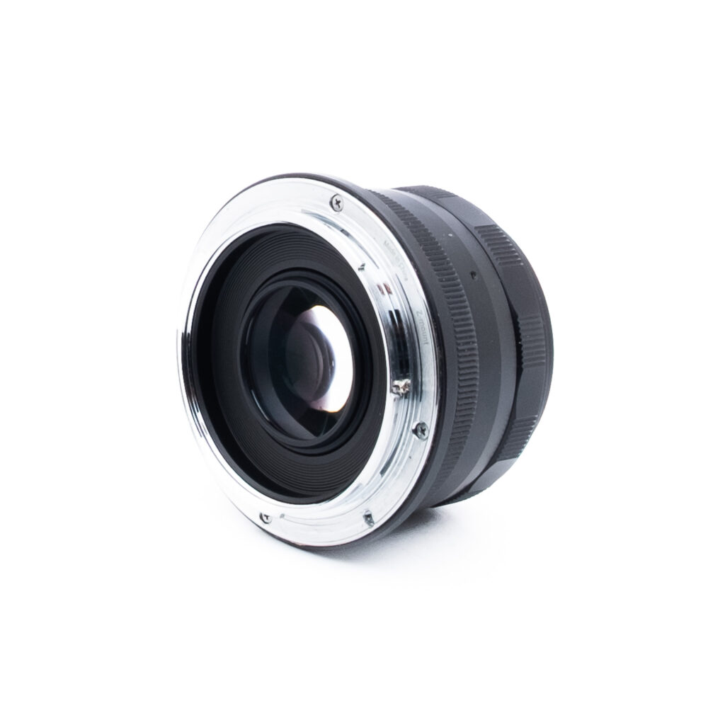Meike 35mm f14 Nikon Z-1 Lähikuva valkoiselle pinnalle asetetusta kameran objektiivista, jossa metallinen kiinnitys ja lasielementit ovat katsojaan päin, ja objektiivin tynnyri ulottuu taaksepäin.