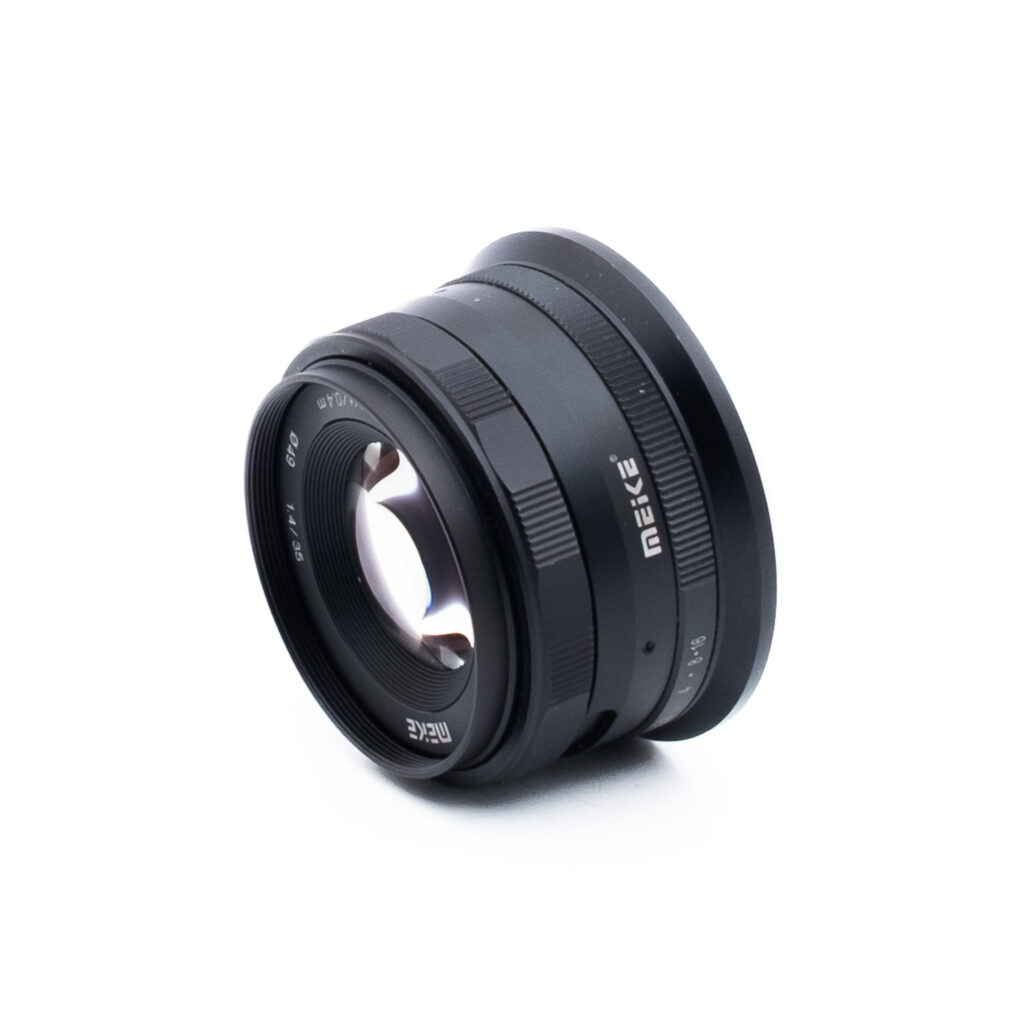 Meike 35mm f14 Nikon Z-2 Musta Meike-kameran objektiivi, jossa on näkyviä lasielementtejä ja kaiverrettuja merkintöjä, on kuvattu kulmassa valkoisella pohjalla.