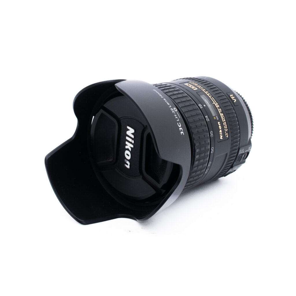 Nikon 16-85mm f35-56G ED DX VR-3 Nikon-kameran objektiivi, johon on kiinnitetty vastavalosuoja, kyljellään valkoista taustaa vasten. Objektiivissa on näkyvät tarkennusrengas ja zoomausrengas, ja siinä on kultaisella ja valkoisella tekstillä merkityt tekniset tiedot.