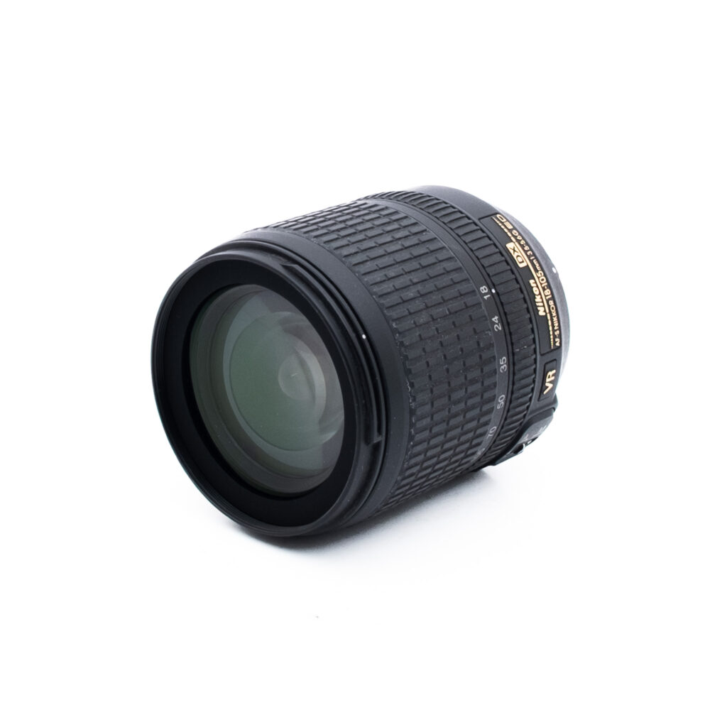 Nikon 18-105mm f35-56 G ED DX VR-2 Musta DSLR-kameran objektiivi, jonka sivulla on kultainen ja valkoinen teksti valkoisella pohjalla ja joka on sijoitettu valkoiselle taustalle niin, että etulasielementti ja kuvioitu tarkennusrengas tulevat esiin.