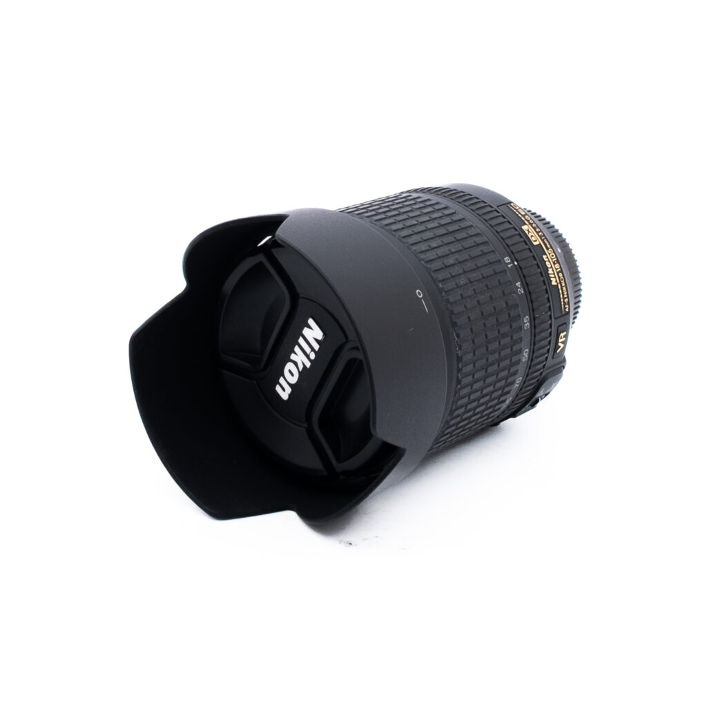 Nikon 18-105mm f35-56 G ED DX VR-3 Nikon-kameran objektiivi, johon on kiinnitetty vastavalosuoja ja objektiivin suojus, valkoisella taustalla.