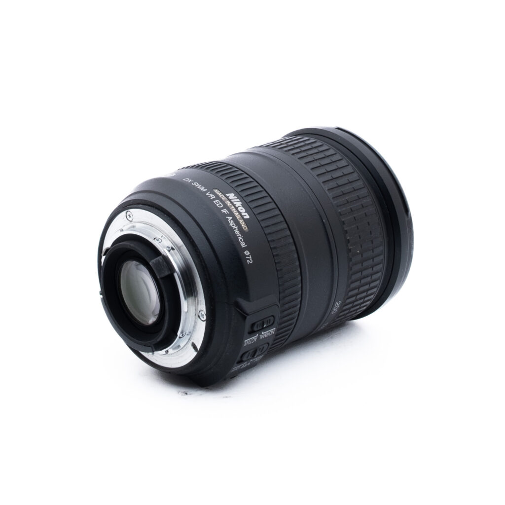Nikon 18-200mm f35-56 G ED DX VR-1 Nikon AF-S DX NIKKOR 18-55mm -kameran objektiivi on kuvassa kyljellään, kiinnitys eteenpäin. Objektiivi on musta, ja siinä on näkyvät tarkennus- ja zoomausrenkaat sekä valkoinen merkintä. Tausta on valkoinen.