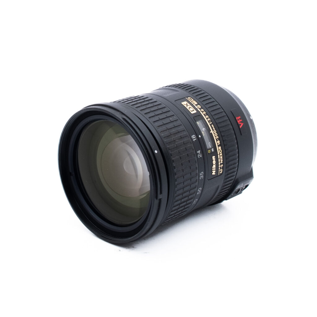 Nikon 18-200mm f35-56 G ED DX VR-2 Musta Nikon-kameran objektiivi, jossa on kultainen ja valkoinen merkintä, näkyvät tarkennus- ja zoomausrenkaat sekä suuri etulasi, valkoisella taustalla.