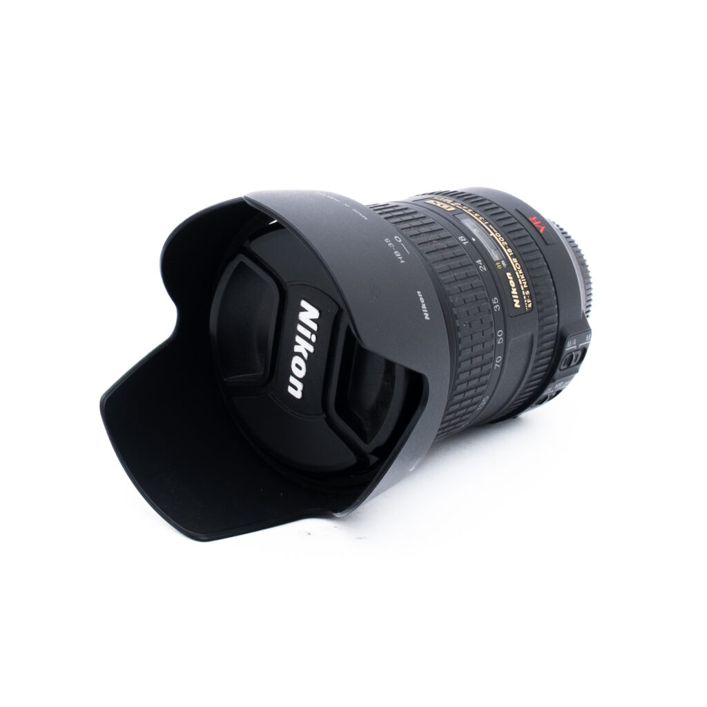 Nikon 18-200mm f35-56 G ED DX VR-3 Nikon-kameran objektiivi, johon on kiinnitetty vastavalosuoja, makaa kyljellään valkoisella taustalla. Objektiivin suojuksessa on näkyvästi Nikonin logo.