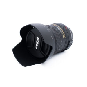 Nikon 18-200mm f35-56 G ED DX VR-3 Nikon-kameran objektiivi, johon on kiinnitetty vastavalosuoja, makaa kyljellään valkoisella taustalla. Objektiivin suojuksessa on näkyvästi Nikonin logo.
