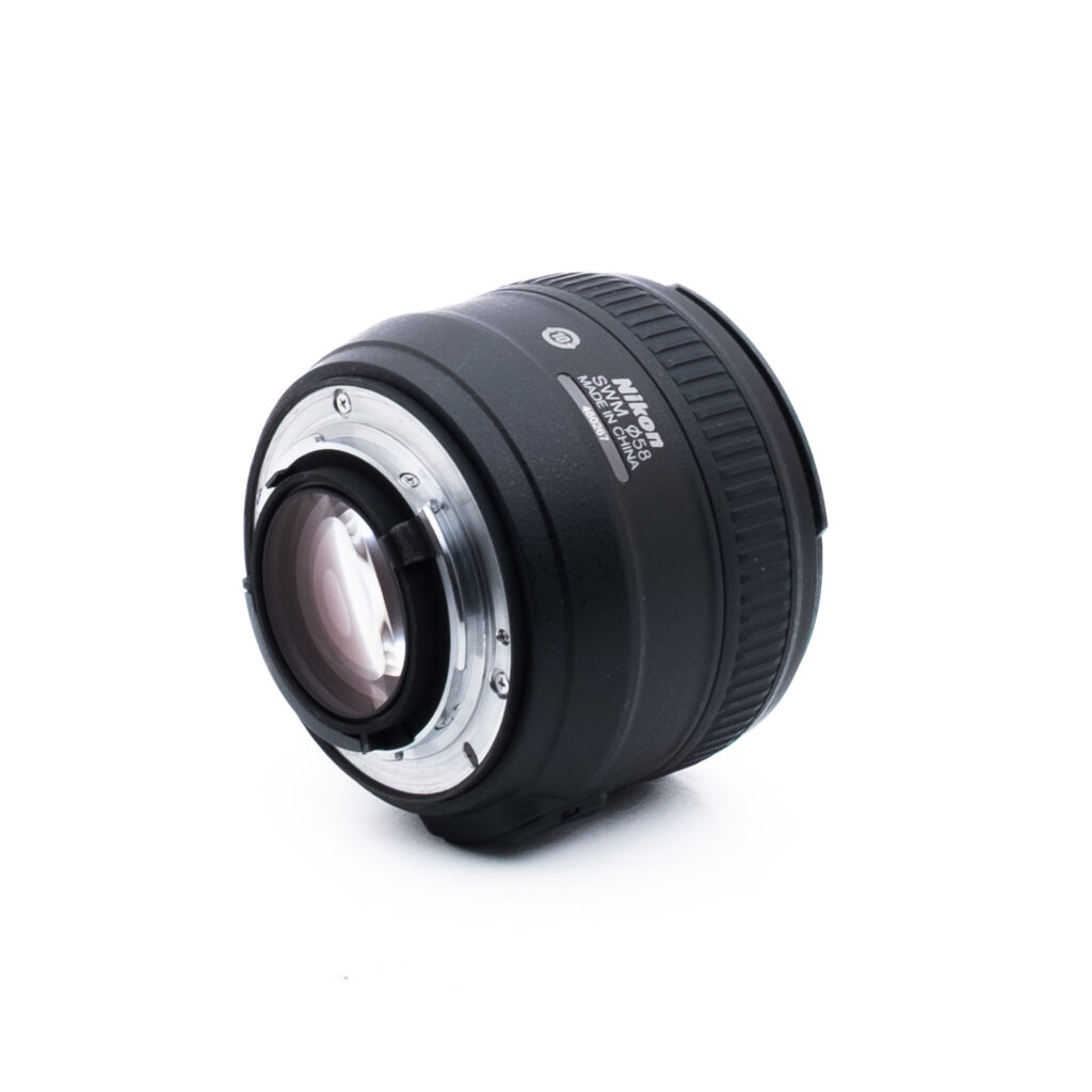 Nikon 50mm f14 G-1-2 Musta Nikon-kameran objektiivi, jossa on hopeinen metallinen kiinnitysrengas, vinossa valkoisella taustalla. Objektiivin merkinnät ja kiinnityskontaktit ovat näkyvissä.