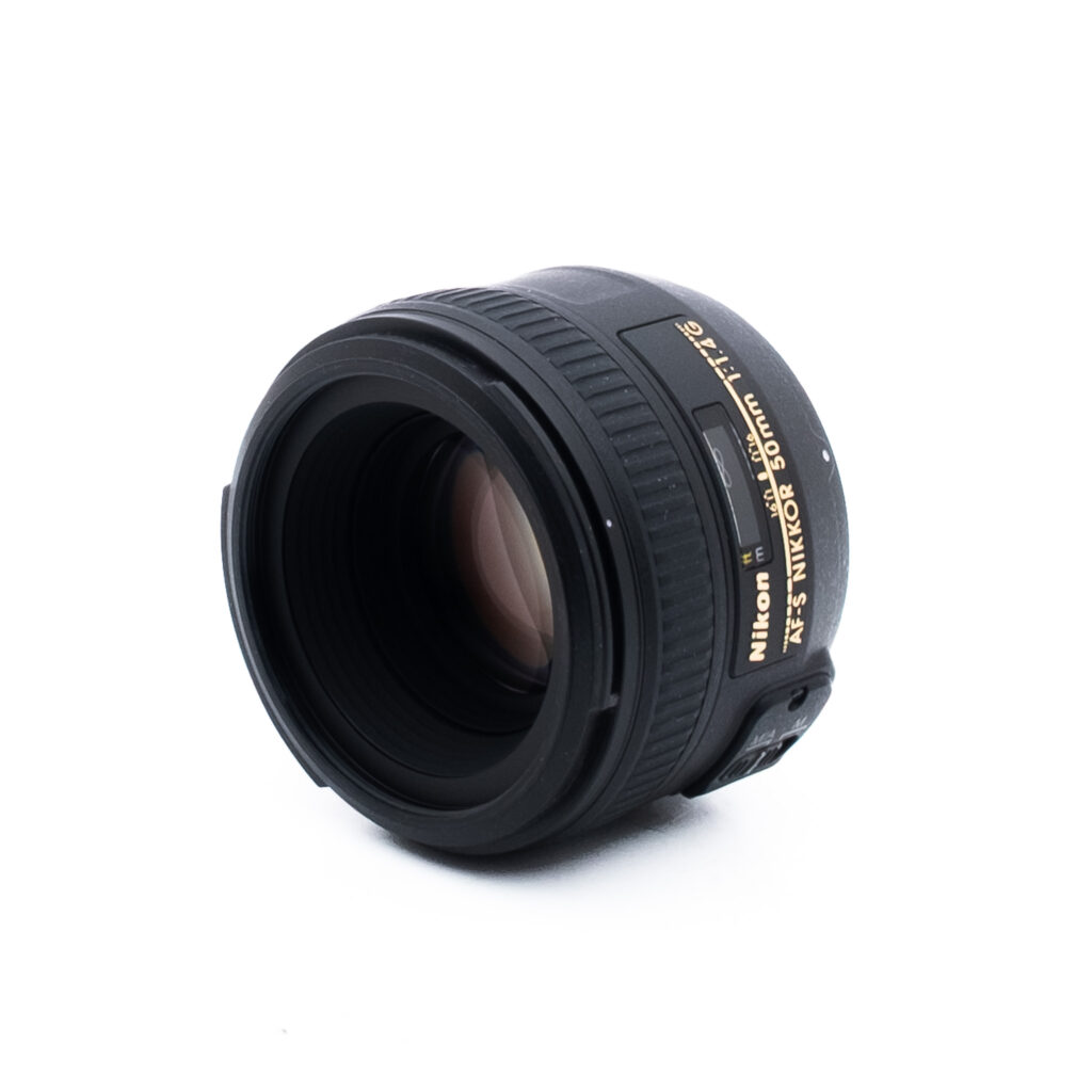 Nikon 50mm f14 G-2-2 Musta Nikon AF-S NIKKOR 50mm f/1.8G -kameran objektiivi valkoisella pohjalla, jossa on kulma, jossa näkyy lasielementti ja kultaiset tuotemerkin yksityiskohdat.