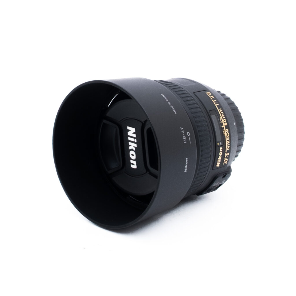 Nikon 50mm f14 G-3-2 Nikon-kameran objektiivi, johon on kiinnitetty vastavalosuoja ja suojus, asetettuna valkoiselle pinnalle. Objektiivissa on kultainen ja valkoinen teksti, joka osoittaa sen tekniset tiedot ja tuotemerkin.