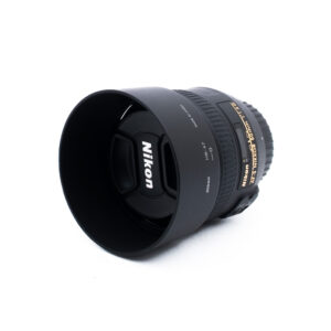 Nikon 50mm f14 G-3-2 Nikon-kameran objektiivi, johon on kiinnitetty vastavalosuoja ja suojus, asetettuna valkoiselle pinnalle. Objektiivissa on kultainen ja valkoinen teksti, joka osoittaa sen tekniset tiedot ja tuotemerkin.