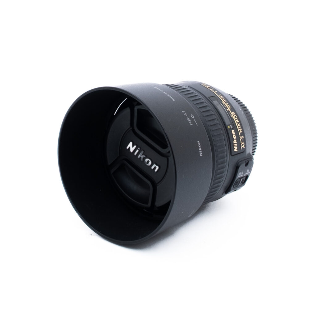Musta Nikon-kameran objektiivi, jossa on vastavalosuoja ja suojus, sekä kultainen ja valkoinen teksti valkoisella pohjalla.