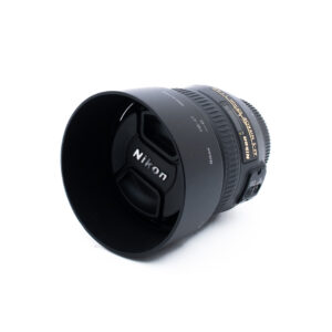 Musta Nikon-kameran objektiivi, jossa on vastavalosuoja ja suojus, sekä kultainen ja valkoinen teksti valkoisella pohjalla.