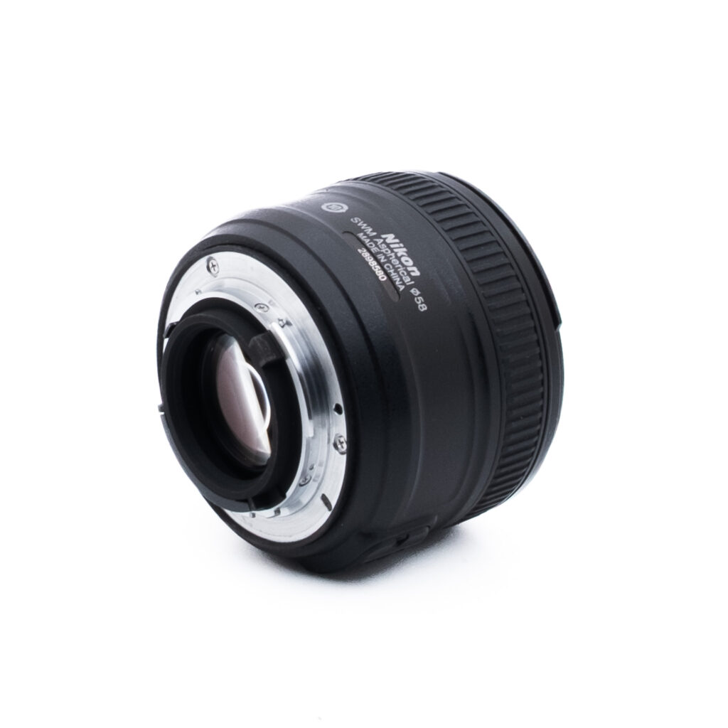 Nikon 50mm f18 G-1-5 Nikon AF-S NIKKOR 50mm f/1.8G -kameran objektiivi näkyy valkoisella pohjalla, ja sen kulma osoittaa metallisen objektiivin kiinnikkeen ja osan mustasta, uurretusta tarkennusrenkaasta.