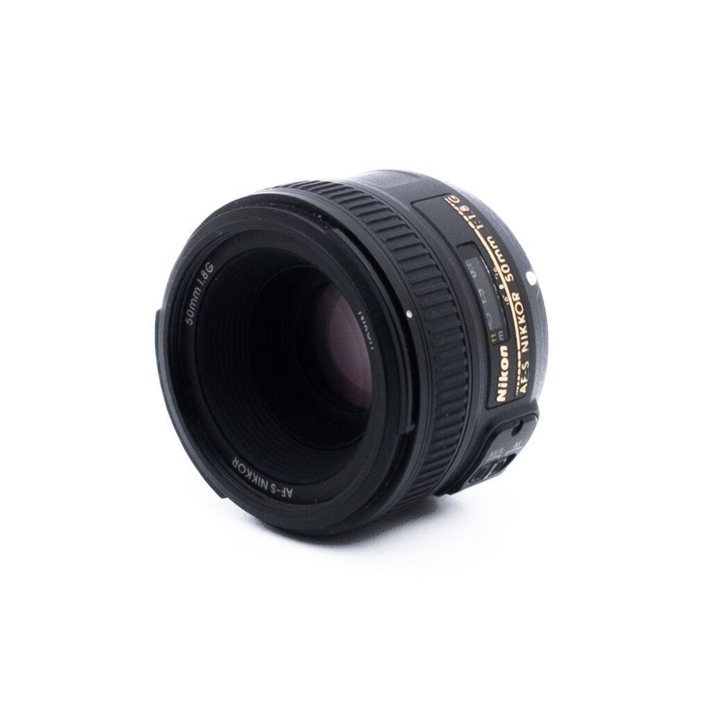 Nikon 50mm f18 G-2-5 Musta Nikon AF-S NIKKOR 50mm f/1.8G -kameran objektiivi on sijoitettu valkoiselle taustalle hieman vasemmalle, ja siinä näkyy objektiivin lasi ja osa kultaisesta tuotemerkinnästä.
