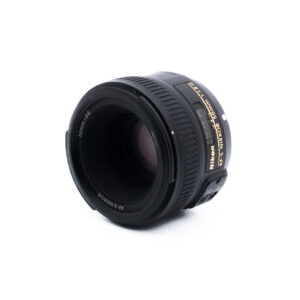 Nikon 50mm f18 G-2-5 Musta Nikon AF-S NIKKOR 50mm f/1.8G -kameran objektiivi on sijoitettu valkoiselle taustalle hieman vasemmalle, ja siinä näkyy objektiivin lasi ja osa kultaisesta tuotemerkinnästä.