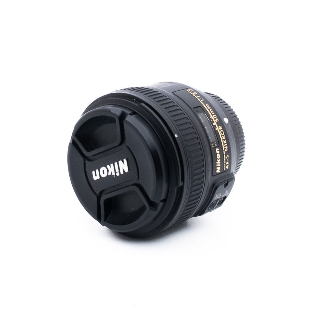 Nikon 50mm f18 G-3-5 Nikon-kameran objektiivi, johon on kiinnitetty objektiivin suojus ja jossa näkyy tuotemerkki ja objektiivin yksityiskohdat valkoisella taustalla.