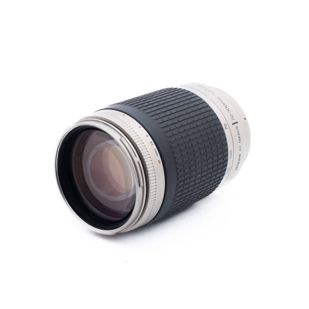 Nikon AF Nikkor 70-300mm f4-56 G-2 Lähikuva kameran zoom-objektiivista, jossa on hopeanvärinen ja musta kuvioitu runko valkoisella pinnalla ja jossa näkyy objektiivin lasi ja osa piipusta.