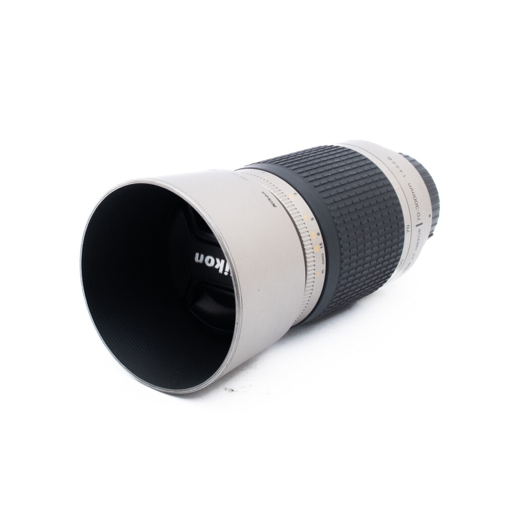 Nikon AF Nikkor 70-300mm f4-56 G-3 Hopeanvärinen ja musta Nikon-kameran objektiivi, jossa on kuvioitu kahva ja pidennetty linssinsuojus ja joka on sijoitettu vinoon valkoiselle taustalle.