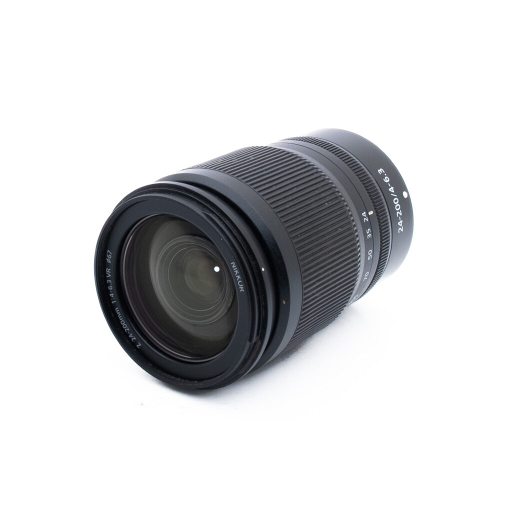 Nikon Z 24-200mm f4-63 VR-2-3 Musta kameran zoom-objektiivi, jossa on valkoiset merkinnät, on sijoitettu valkoiselle pinnalle, ja se on taivutettu niin, että lasinen etupaneeli ja kuvioitu zoom-rengas tulevat näkyviin.