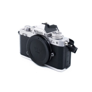 Nikon Z fc-5 Hopeanmusta Nikon Z fc (SC 6500) - Käytetty peilitön kamera on kuvassa vasemmalle päin, runkokorkki päällä, ja siinä on retrovaikutteiset valintakiekot ja kuvioitu kahva valkoista taustaa vasten. Ei objektiivia kiinnitettynä.