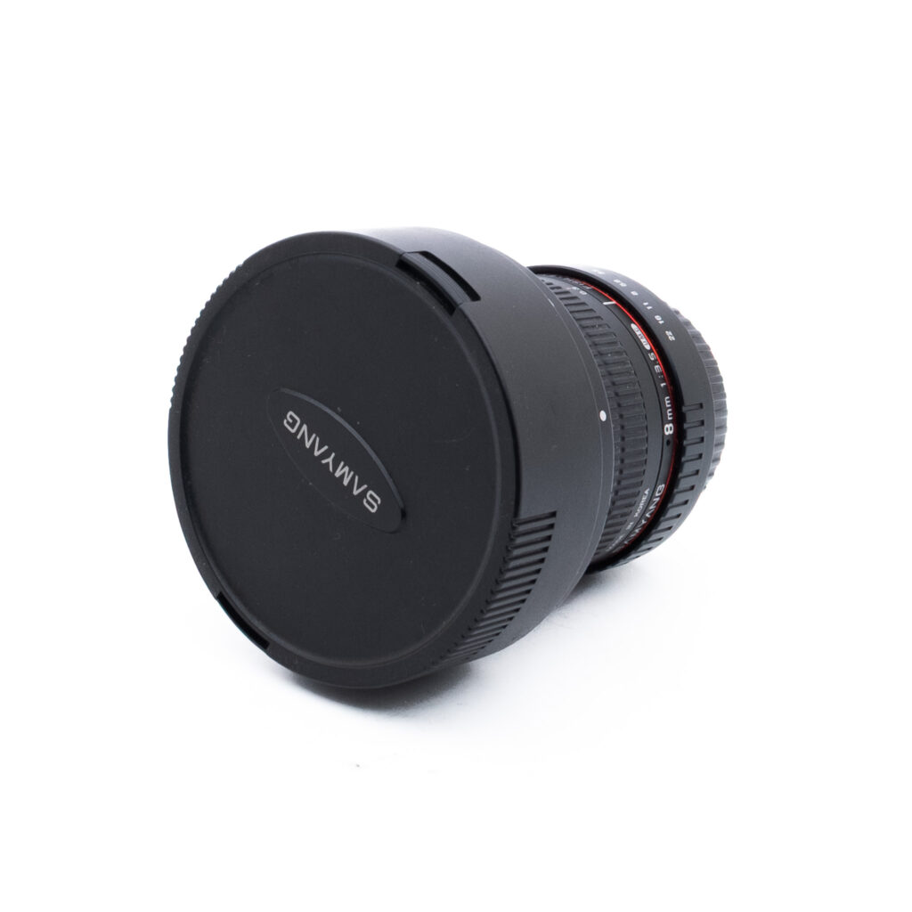 Samyang 8mm f35 UMC-3-2 Mustalla suojuksella varustettu kameran objektiivi, jossa on merkintä Samyang, on kuvattu kulmassa valkoisella taustalla. Objektiivissa on harjatut tarkennusrenkaat ja aukkorenkaat, joissa on punaiset ja valkoiset merkinnät.