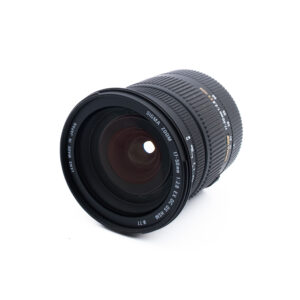 Sigma 17-50mm f28 EX DC OS HSM Canon-2-2 Musta Sigma 17-50 mm:n kameran objektiivi, jossa on valkoinen teksti, jossa ilmoitetaan tekniset tiedot. Objektiivi on sijoitettu valkoiselle taustalle, ja sitä on taivutettu niin, että etulasi ja osa objektiivin tynnyristä näkyvät.