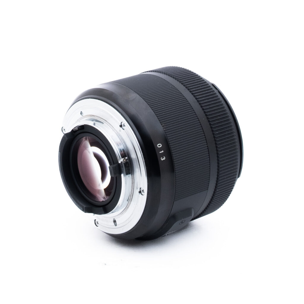 Sigma 30mm f14 DC Nikon-1 Lähikuva mustasta kameran objektiivista takakiinnityspuolelta katsottuna, jossa näkyy metallinen kiinnitysrengas ja lasielementit valkoista taustaa vasten.