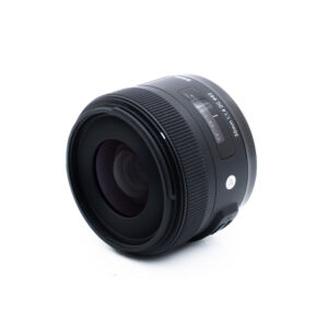 Sigma 30mm f14 DC Nikon-2 Musta kameran linssi, jossa on näkyvä tarkennus ja automaattinen/manuaalinen kytkin, asetettuna valkoiselle taustalle ja kulmattuna siten, että etulasi ja osa piipun yksityiskohdista tulevat näkyviin.