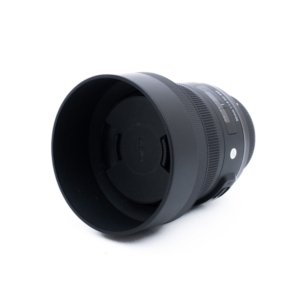 Sigma 30mm f14 DC Nikon-3 Musta Sigma-kameran objektiivi, johon on kiinnitetty vastavalosuoja ja suojus, valkoisella taustalla, hieman vasemmalle kallistettuna.