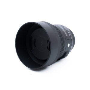 Sigma 30mm f14 DC Nikon-3 Musta Sigma-kameran objektiivi, johon on kiinnitetty vastavalosuoja ja suojus, valkoisella taustalla, hieman vasemmalle kallistettuna.