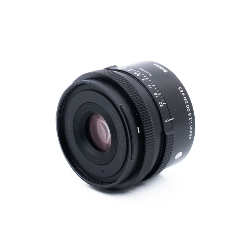 Sigma 45mm f28 DG DN Sony-2 Musta kameran objektiivi, jossa on valkoinen teksti, mukaan lukien tiedot 45mm 1:2.8 DG DN ja Sigma, sijoitettu valkoiselle taustalle, vinossa niin, että etulasi ja sivumerkinnät näkyvät.