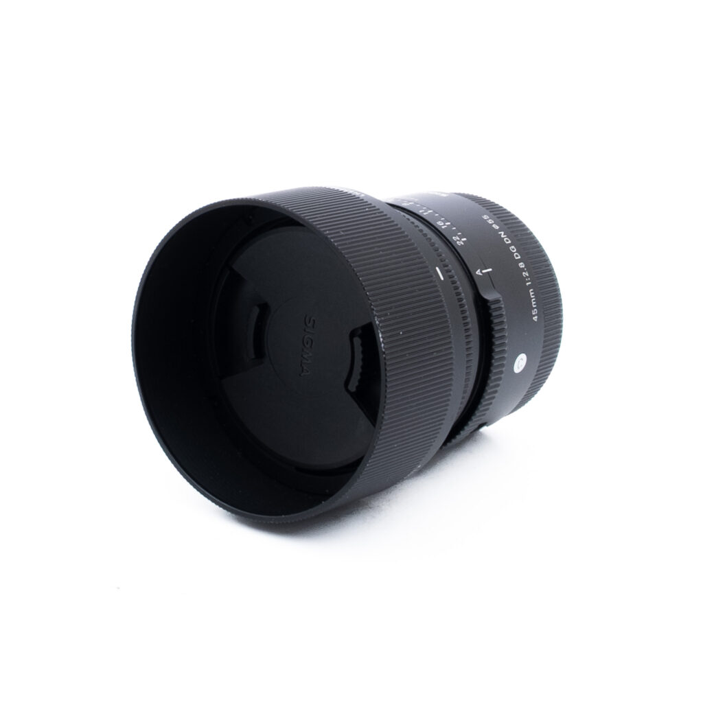 Sigma 45mm f28 DG DN Sony-3 Musta Sigma-kameran objektiivi, johon on kiinnitetty linssinsuojus, makaa valkoisella pinnalla ja on hieman oikealle kallistettu. Objektiivissa on näkyvät harjatut tarkennusrenkaat ja tuotemerkit.