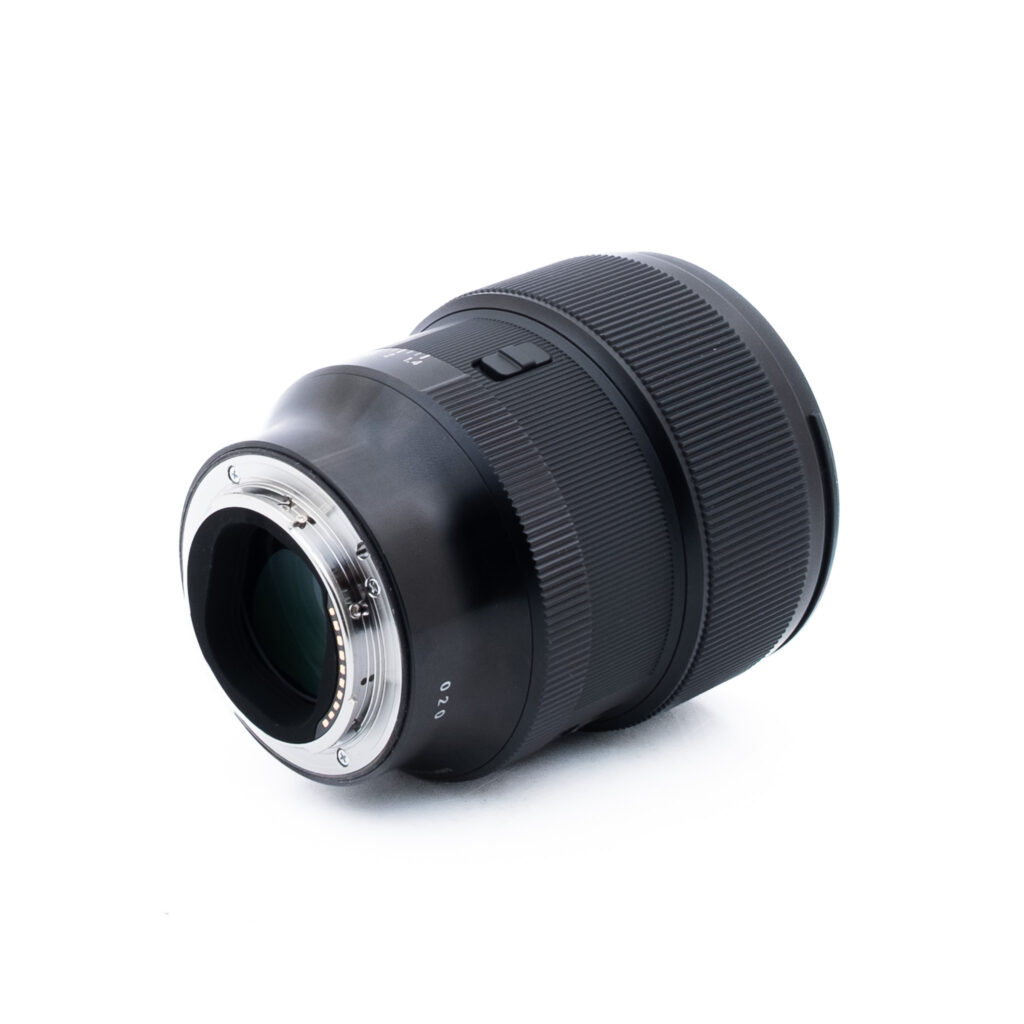 Sigma 85mm f14 DG DN Sony-1-4 Musta kameran objektiivi, jossa on metallinen kiinnitysrengas, on asetettu kyljelleen valkoista taustaa vasten. Objektiivissa on kuvioidut pitorenkaat ja näkyvät elektroniset koskettimet kiinnityspäässä.