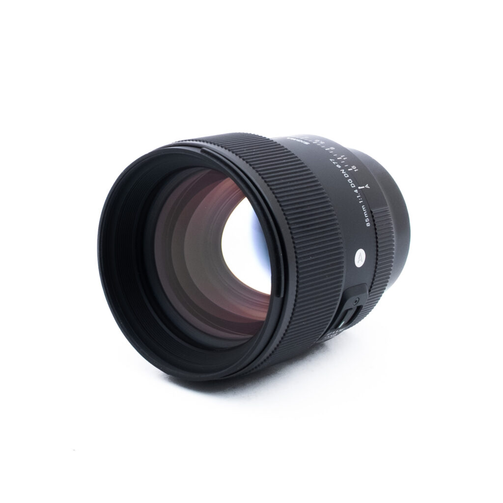 Sigma 85mm f14 DG DN Sony-2-4 Musta kameran objektiivi, jossa on uurrettu tarkennusrengas ja näkyvät lasielementit, on sijoitettu vinoon valkoiselle taustalle. Objektiivin rungossa on valkoinen teksti ja hopeinen painike.