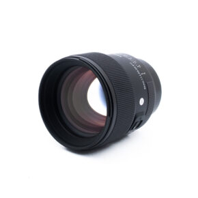 Sigma 85mm f14 DG DN Sony-2-4 Musta kameran objektiivi, jossa on uurrettu tarkennusrengas ja näkyvät lasielementit, on sijoitettu vinoon valkoiselle taustalle. Objektiivin rungossa on valkoinen teksti ja hopeinen painike.