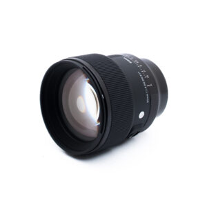 Sigma 85mm f14 DG DN Sony-2-5 Valkoisella taustalla näkyy musta kameran linssi, jossa on suuri lasielementti. Objektiivissa on harjanteiset tarkennusrenkaat ja sivussa näkyvät tuotemerkit.