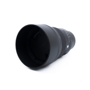Sigma 85mm f14 DG DN Sony-3-4 Musta kameran objektiivi, johon on kiinnitetty suuri linssinsuojus, kuvattuna valkoisella taustalla. Objektiivin suojuksessa näkyy tuotemerkki Sigma.