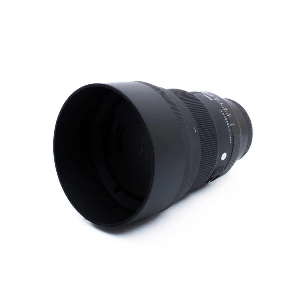 Sigma 85mm f14 DG DN Sony-3-5 Musta kameran objektiivi, johon on kiinnitetty leveä linssinsuojus, on sijoitettu valkoiselle taustalle ja suunnattu hieman oikealle.